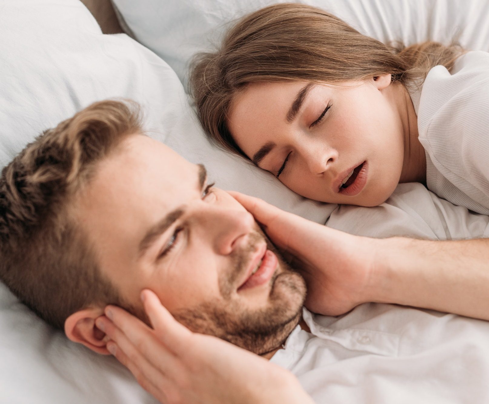 exhausted man plugging ears with hands while lying in bed near snoring wife